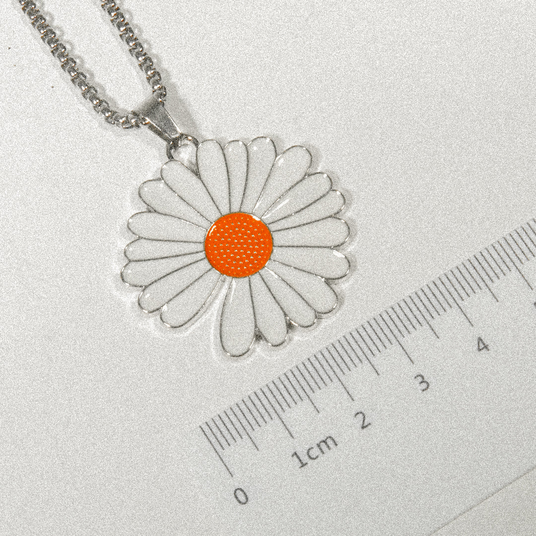 A white daisy pendant featuring an orange center is displayed on a ruler, highlighting its delicate design and size.
