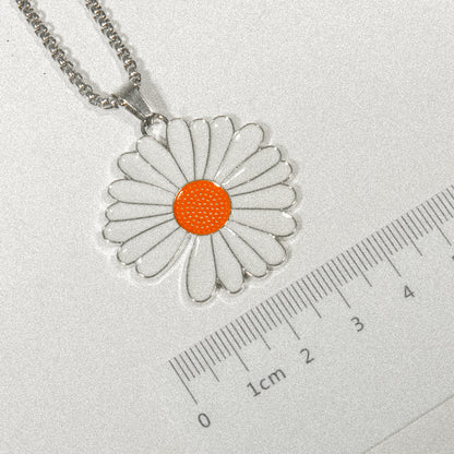 A white daisy pendant featuring an orange center is displayed on a ruler, highlighting its delicate design and size.
