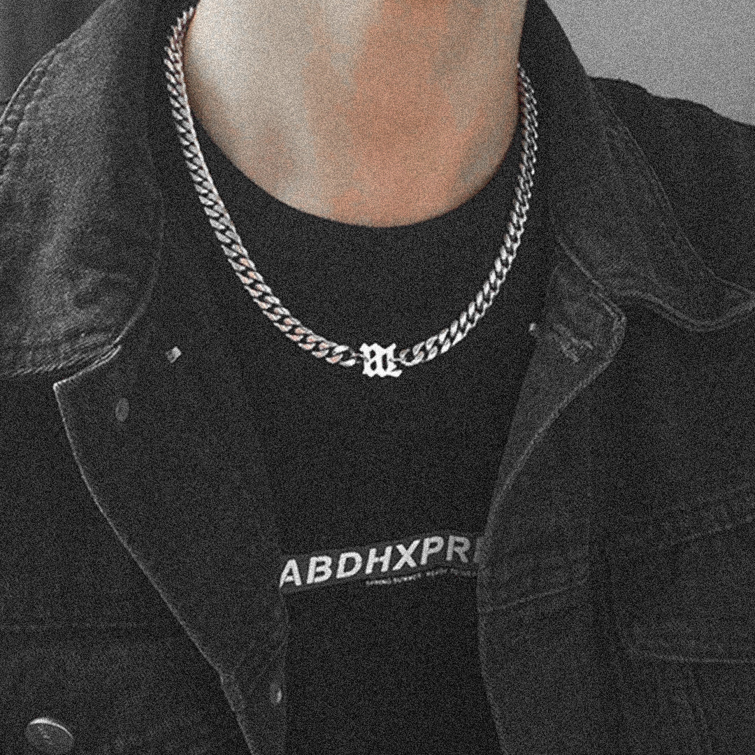 Close-up of a stylish unisex silver chain necklace featuring a bold Gothic letter pendant, worn with a black denim jacket.