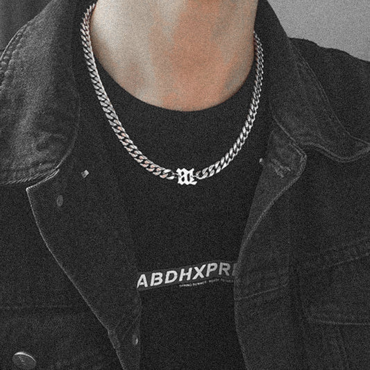 Close-up of a stylish unisex silver chain necklace featuring a bold Gothic letter pendant, worn with a black denim jacket.