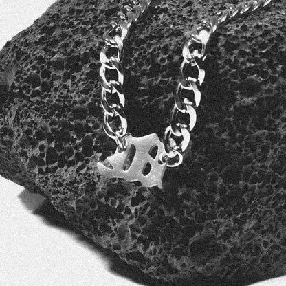 Unique Gothic letter pendant on a silver unisex chain necklace displayed on a textured black stone.
